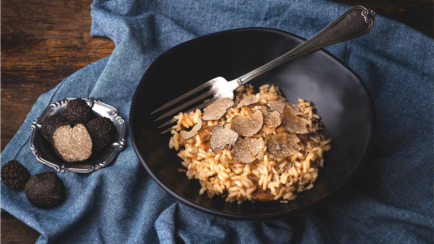 A black bowl of creamy risotto topped with thinly shaved black truffles, served with a silver fork and fresh whole truffles on the side.