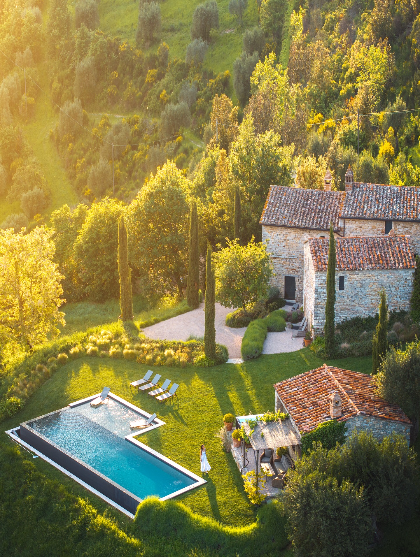Aerial view of a stone villa at Tenuta di Murlo in Umbria, surrounded by olive groves and cypress trees, with a pool glowing in the golden sunset. Photo: @tenutadimurlo