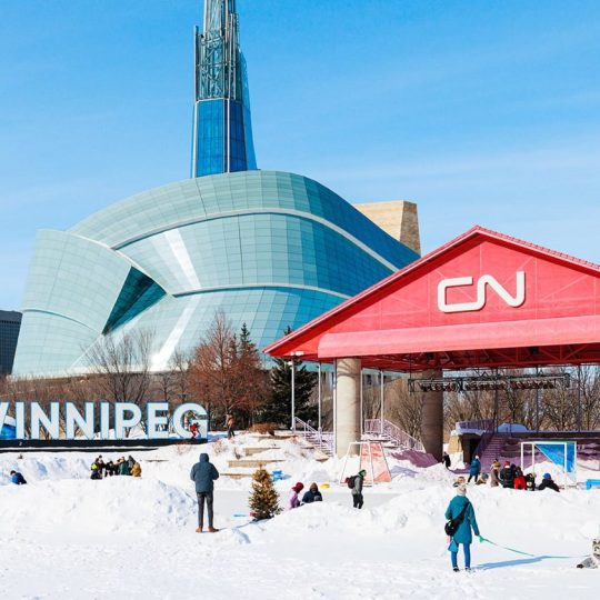 View of Winnipeg skyline from The Forks National Historic Site, showing rivers, historic buildings, and downtown cityscape.