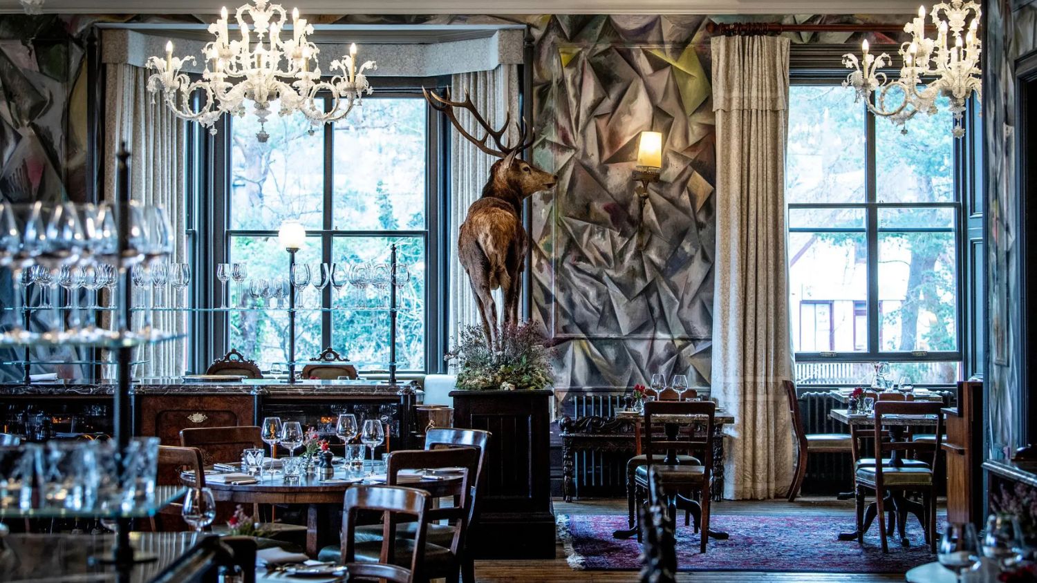 The Clunie Dining Room at the Fife Arms in Braemar, Scotland. The walls feature a kaleidoscopic mural by Argentinian artist Guillermo Kuitca, titled "The Clunie River Mural." A mounted stag brings rustic warmth, while Murano glass chandeliers and dark wood add quiet elegance